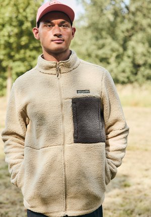 Giacca in pile beige con una patch grigia sul lato sinistro del petto, chiusura con zip, colletto alto e tessuto testurizzato. Abbigliamento casual per attività all'aperto.