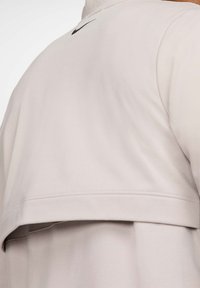 Béžová vrstvená bunda so zatepleným chrbtom a malým čiernym logom Nike pri golieri.