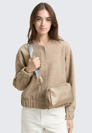 Beiger Strickpullover mit Knopfverschluss, elastischen Bündchen, getragen mit einer metallisch goldenen Umhängetasche mit einem gemusterten Tragegurt in Blau und Beige.
