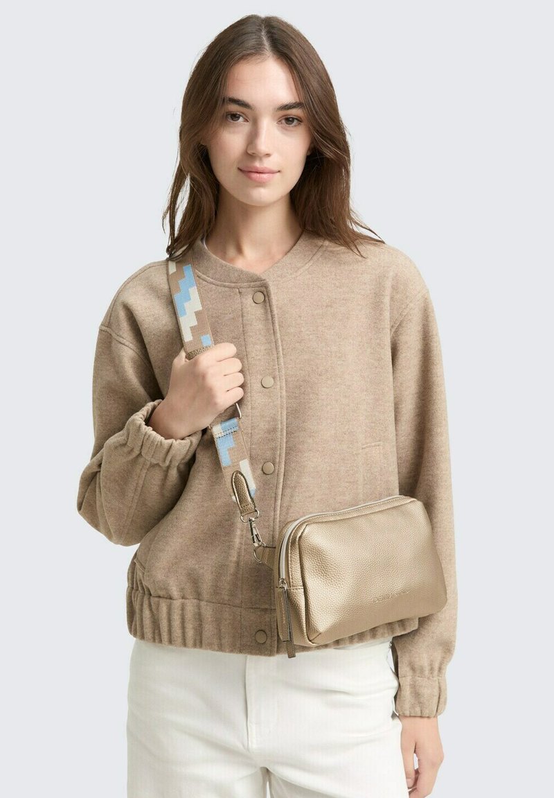 Beiger Strickpullover mit Knopfverschluss, elastischen Bündchen, getragen mit einer metallisch goldenen Umhängetasche mit einem gemusterten Tragegurt in Blau und Beige.