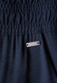 Mörkblått tyg med pucklad textur, prytt med en silverfärgad metalltagg märkta "CECIL." Materialet ser slätt och lättviktigt ut.