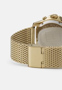Bracelet de montre en maillage doré avec une finition texturée et une boucle de verrouillage. Boîtier de montre rectangulaire avec un dos transparent affichant les détails internes.