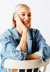 Giacca di jeans con maniche arrotolate e colore blu sbiadito, abbinata a un braccialetto di perline multicolore, seduta su una sedia bianca.