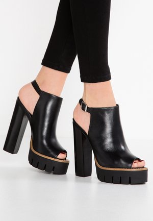 Des chaussures à talons hauts noires avec un fini cuir brillant, présentant une plateforme épaisse et une sangle arrière avec une boucle réglable.