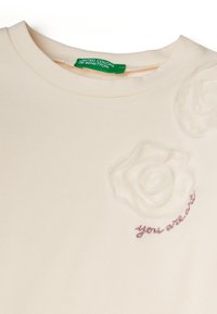 Krémové tričko s kulatým výstřihem, které má vyvýšený květinový vzor a růžový vyšitý text "ty nejsi." Vyrobeno z měkké bavlny.
