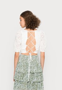 Top de algodão branco com mangas bufantes, detalhes em broderie anglaise e costas em laço cruzado. Combinado com uma saia verde fluida e estampada.