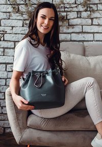 Šedá kožená kabelka ve tvaru kbelíku se stříbrnými detaily, stažením na šňůrku, lesklým povrchem a vyraženým logem na přední straně, kterou drží sedící modelka.