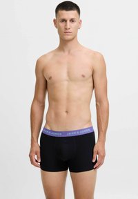 Mlad moški stoji z rokama sproščenima, oblečen v črne boksarice s purpurnim pasom z oznako "JACK & JONES," na enostavni svetli ozadju.