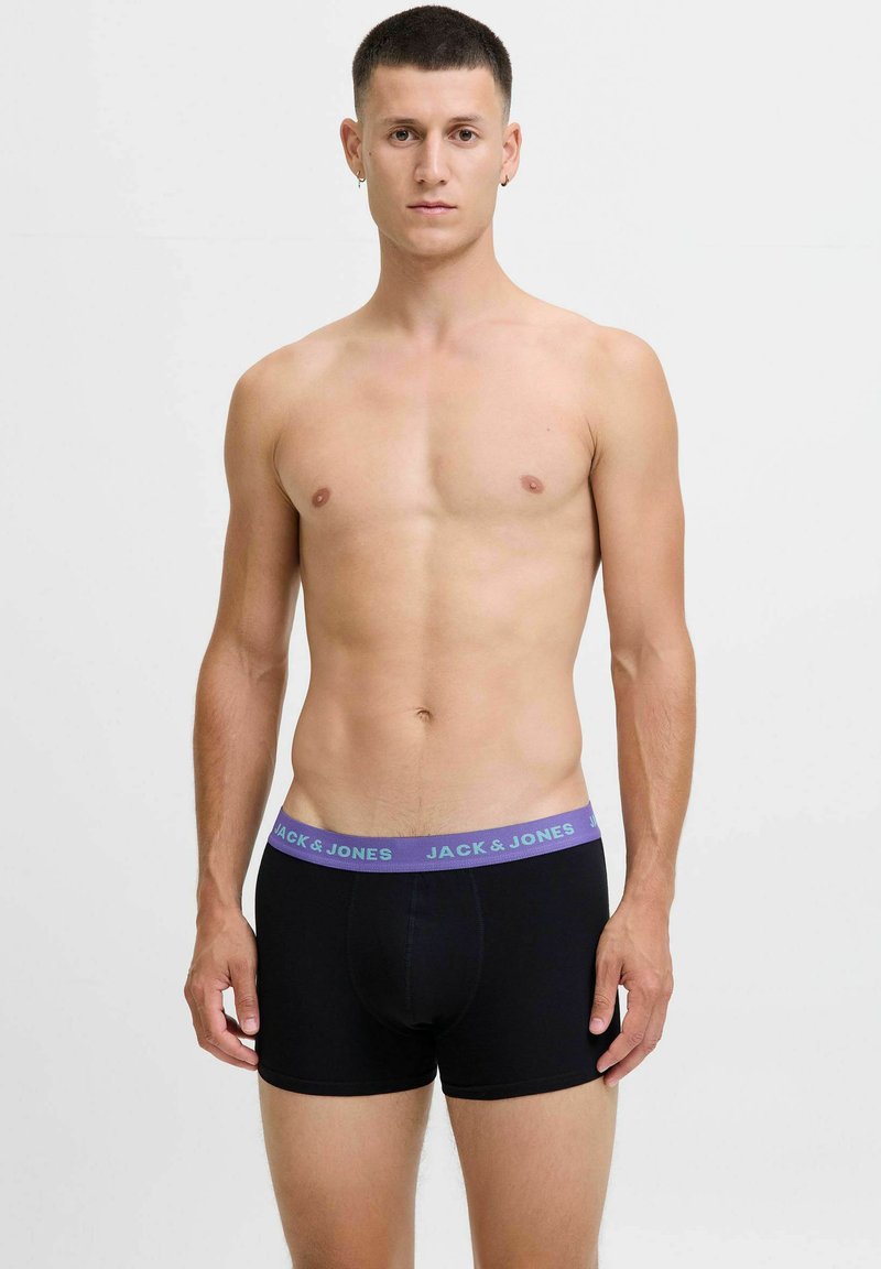 Mlad moški stoji z rokama sproščenima, oblečen v črne boksarice s purpurnim pasom z oznako "JACK & JONES," na enostavni svetli ozadju.