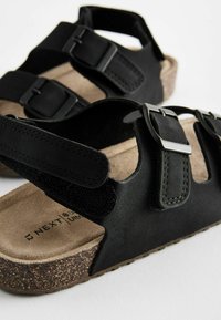 Svarta sandaler med två justerbara spännen, tillverkade av släta och texturerade material. Korkliknande sula med en mjuk innersula.