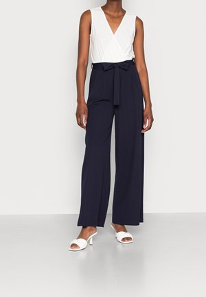 Kobieta w bezrękawniku z białej, kopertowej bluzce, granatowych, szerokich spodniach z wysokim stanem i materiałowym paskiem oraz białych sandałach na obcasie z odkrytymi palcami stoi na szarej podłodze.