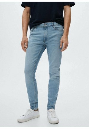 Mann trägt hellblaue Slim-Fit-Jeans, ein schwarzes T-Shirt und weiße Sneakers, steht vor einem schlichten weißen Hintergrund.