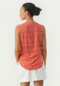 Rote ärmellose Bluse mit einem weißen abstrakten Muster, rundem Saum und weichem Stoff. Das Modell wird von hinten gezeigt, kombiniert mit einem weißen Rock.