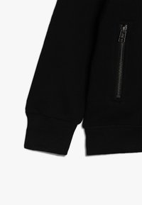 Suda de color negro con una bolsa con cremallera en la manga izquierda, puños acanalados y un diseño ajustado. Textura suave con un detalle mínimo.