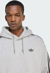 Sivá mikina s kapucňou a sťahovacou šnúrkou, s čiernymi trojpásikovými detailmi a malým čiernym logom na hrudi. Z materiálu z bavlnenej zmesi.