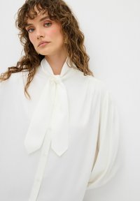 Blouse blanche à manches longues et nœud cou. Fabriquée en tissu léger, elle présente un boutonnage à l'avant et un discret plissement au niveau des épaules.