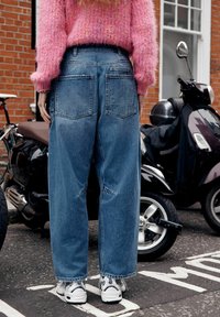 Jasnoniebieskie, szerokie dżinsy z wysokim stanem, postrzępionymi mankietami i dwoma tylnymi kieszeniami, zestawione z białymi trampkami. Tło z ceglaną ścianą.