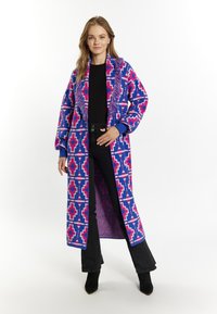 Cardigan lung, cu un model în albastru și roz, având un guler cu franjuri și deschidere în față, asortat cu un top și pantaloni negri. Botine cu toc negre.