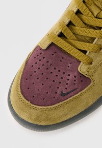 Calçado Nike em camurça amarelo-verde com a biqueira bordô. Os detalhes notáveis incluem perfurações, logótipo preto e atacadores texturizados.