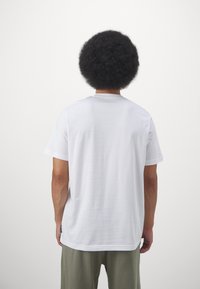 Bărbat cu coafură afro, purtând un tricou alb simplu cu mânecă scurtă și pantaloni verzi măslinii, stând cu spatele pe un fundal alb.