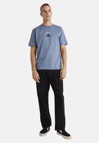 Camiseta de algodón azul claro con mangas cortas y escote redondo, que presenta un pequeño gráfico en el pecho; combinada con pantalones negros de corte relajado.