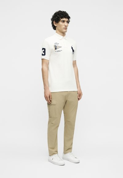 Άνδρας που στέκεται φορώντας λευκό πόλο με σκούρο μπλε λογότυπο και σημάδια, μπεζ παντελόνια cargo και λευκά αθλητικά παπούτσια σε απλό φόντο.