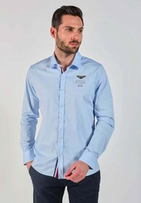 Chemise à manches longues de couleur bleu clair avec un col boutonné. Présente un logo brodé et une patte de boutonnage intérieure contrastée avec des rayures rouges et blanches.