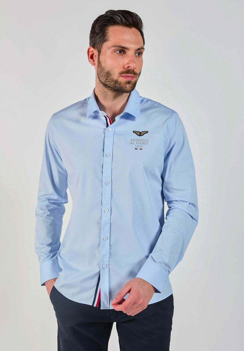 Chemise à manches longues de couleur bleu clair avec un col boutonné. Présente un logo brodé et une patte de boutonnage intérieure contrastée avec des rayures rouges et blanches.