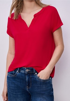Kvinde iført en lys rød kortærmet top med V-hals kombineret med blå denimjeans, den ene hånd i lommen, foran en ensfarvet baggrund.