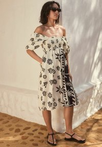 Off-the-shoulder, beige klänning med svarta blommönster, smockad midja och knälång fåll. Bärs med svarta flip-flops. Slät textur.