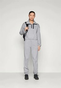 Mulher em pé, vestindo um fato de treino cinza e sapatilhas pretas, segurando a alça de uma mochila preta com uma expressão neutra, em um fundo claro e simples.