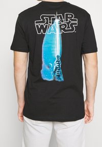 Musta puuvillapaita, jossa on suuri sininen ja valkoinen graafinen kuva valosabasta ja "STAR WARS" -teksti takana. Lyhyet hihat ja pyöreä kaula-aukko.
