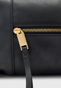 Gros plan sur un sac en cuir noir avec une fermeture éclair dorée et une languette en cuir attachée au tirette de la fermeture.