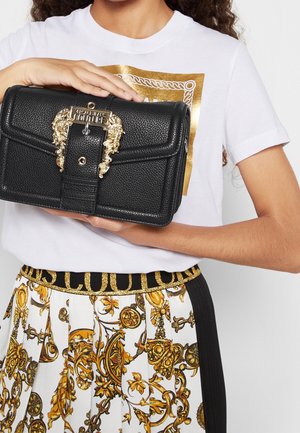 Žena držící černou texturovanou kabelku se zdobenou zlatou sponou, oblečená v bílém tričku a bílé sukni se zlatým barokním potiskem a černým pasem.