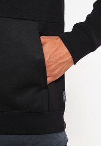 Felpa nera con un davanti texturizzato, tasca a marsupio e polsini a costine. Il tessuto appare morbido, con un'etichetta visibile sulla cucitura laterale.