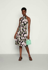 Mulher vestindo um vestido preto sem mangas com estampado floral em rosa e verde, segurando uma pequena bolsa verde menta, de pé contra um fundo liso.