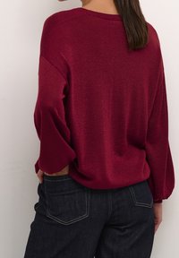 Pull bordeaux avec de longues manches bouffantes et un col rond, fabriqué en tissu tricoté texturé. Associé à un jean denim bleu foncé.