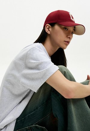 Burgundská čiapka s béžovým okrajom, šedé tričko s krátkym rukávom a zelené rifle. Postava sedí, pričom je dôraz na neformálnom oblečení.