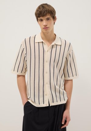 Junge Person trägt ein cremefarbenes kurzärmeliges Strickshirt mit schwarzen vertikalen Streifen und schwarze Hose, steht vor einem schlichten hellen Hintergrund.