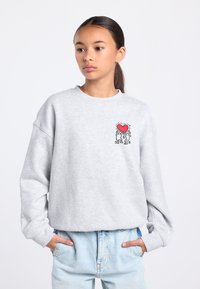 Sweatshirt gris clair avec un col rond, des manches oversize et un graphique sur la poche avant représentant un cœur rouge. Tissu doux.