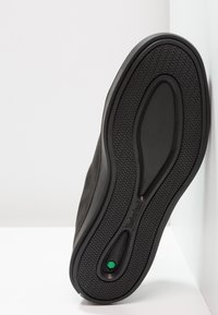 Semelle de chaussure noire avec un motif ondulé texturé et un détail hexagonal vert près du talon, positionnée verticalement contre une surface blanche.