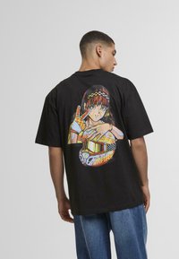 Svart oversized t-skjorte med et fargerikt anime-inspirert grafikk på baksiden av en karakter med hjelm og hansker, mot en ensfarget bakgrunn.