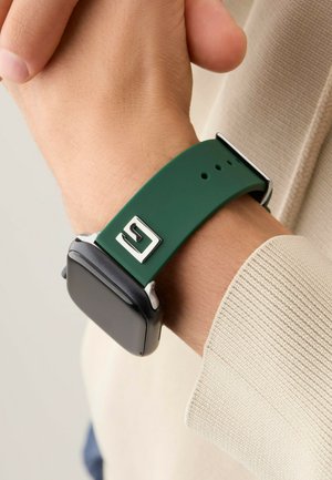 Correa de reloj de silicona verde con un acento cuadrado plateado y una esfera cuadrada negra, llevada sobre un suéter beige claro.