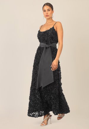 Schwarzes Abendkleid aus floraler Spitze mit enganliegendem Oberteil, Spaghettiträgern und einem fließenden Rock. Mit einem Satin-Gürtel, der in der Taille gebunden ist. Silberne Absätze.