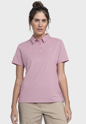 Frau mit dunklen Haaren, zu einem Dutt zusammengebunden, trägt ein hellviolettes Poloshirt mit kurzen Ärmeln und beige Hose, steht vor einem einfarbigen hellen Hintergrund.