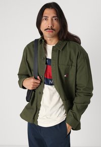 Chemise verte à boutons en coton, dotée d'une poche poitrine et d'un petit écusson logo. Portée par-dessus un t-shirt graphique de couleur claire.
