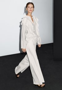White ruffled blouse with a V-neck and drawstring waist, paired with wide-leg white pants. The model wears brown platform sandals.
