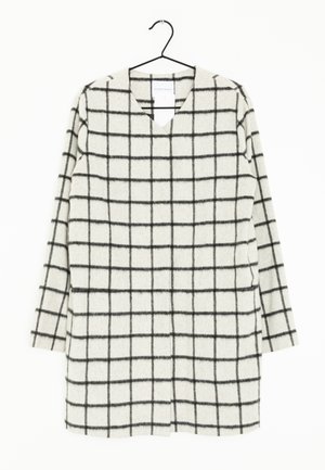 Wit jurk met lange mouwen en zwart ruitpatroon, V-hals, hangend aan een zwarte hanger tegen een witte achtergrond.