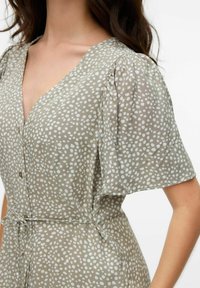 Robe légère olive avec des pois blancs, décolleté en V profond, détails de boutons et manches bouffantes, cintrée à la taille par un lien.