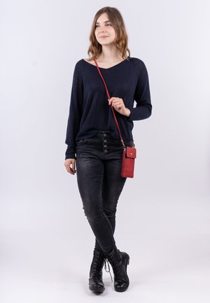 Marineblaues Strickoberteil, schwarze enge Jeans und schwarze Schnürstiefel. Eine rote Umhängetasche für das Handy hängt über ihrer Schulter. Neutraler Hintergrund.